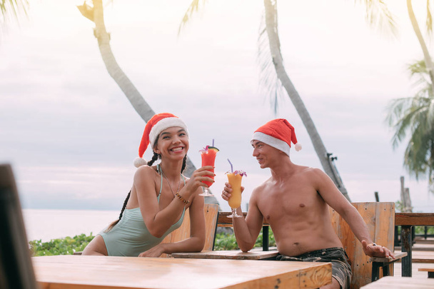 Genç bir çift, tropik bir adada Noel şapkası takıp okyanusta kokteyl içiyor..  - Fotoğraf, Görsel