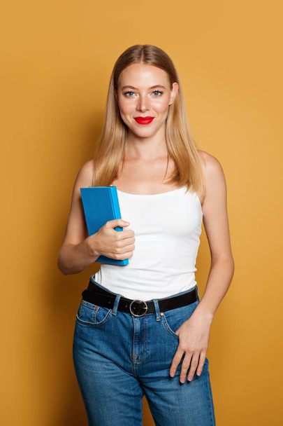Young woman student holding blue book on bright yellow background - 写真・画像
