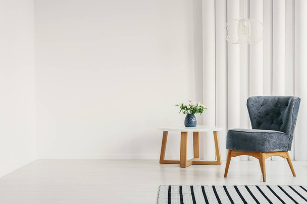Fashionable blue armchair next to white round coffee table with vase with flowers in empty white interior with striped rug, real photo with copy space on empty white wall - Foto, afbeelding