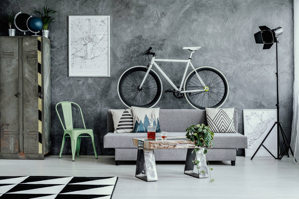 Mint green metal chair next to grey sofa with pillow in dark living room interior with map on empty concrete wall and industrial furniture - Фото, изображение