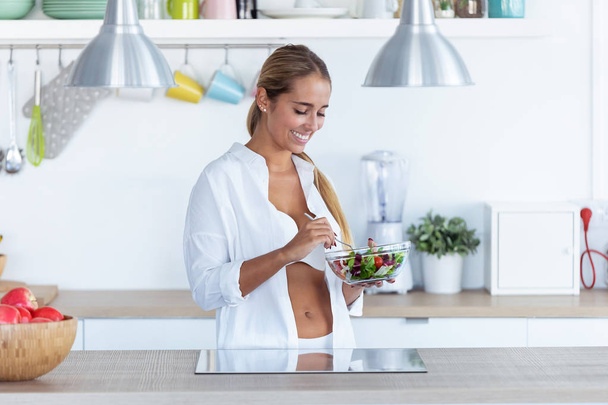 Csinos fiatal nő fehérneműben salátát eszik otthon a konyhában.. - Fotó, kép