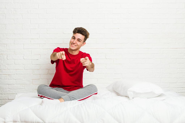 młody nastolatek student człowiek na łóżku wesoły uśmiech wskazując do przodu. - Zdjęcie, obraz