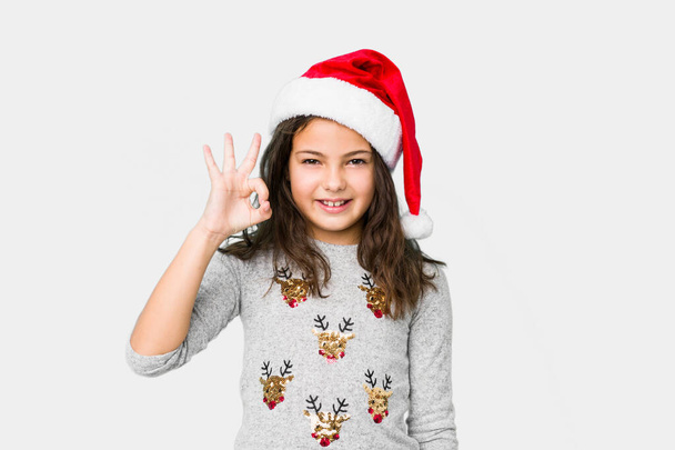 Little girl celebrating christmas day winks an eye and holds an okay gesture with hand. - Fotó, kép