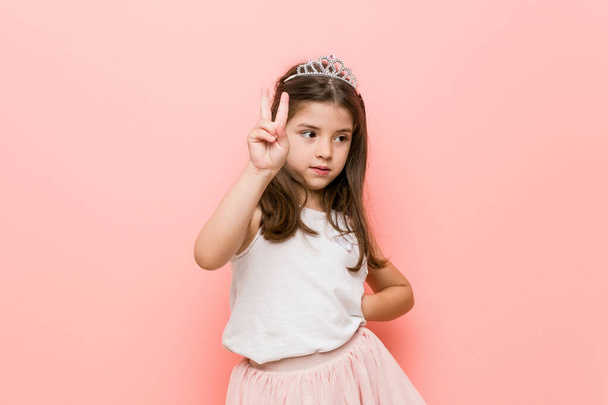 Маленька дівчинка в принцесі виглядає радісно і безтурботно, показуючи символ миру з пальцями
. - Фото, зображення