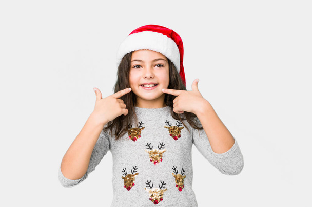 Little girl celebrating christmas day smiles, pointing fingers at mouth. - 写真・画像