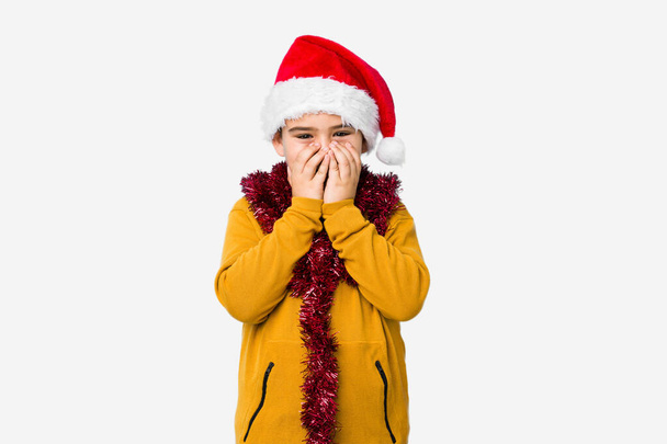 Ένα μικρό αγόρι γιορτάζει τα Χριστούγεννα φορώντας ένα καπέλο Σάντα απομονωμένο γελώντας για κάτι, καλύπτοντας το στόμα με τα χέρια. - Φωτογραφία, εικόνα