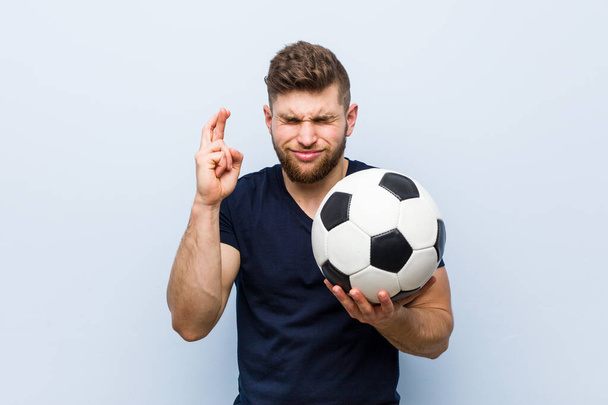Young caucasian man holding a soccer ball crossing fingers for having luck - Photo, Image