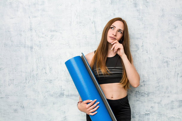 Young caucasian sportswoman holding a mat looking sideways with doubtful and skeptical expression. - Photo, Image