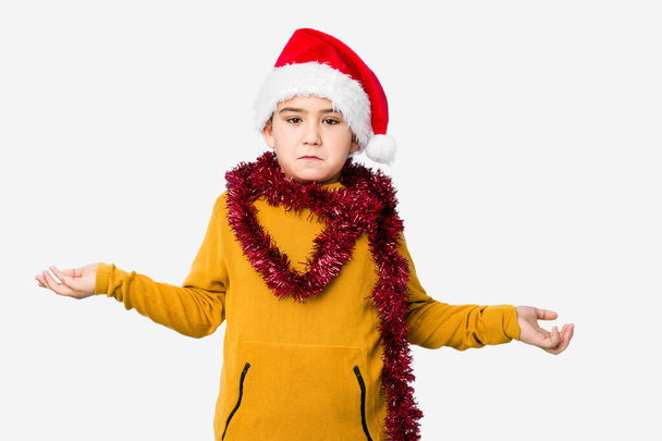 Little boy celebrating christmas day wearing a santa hat isolated confused and doubtful shrugging shoulders to hold a copy space. - Фото, зображення