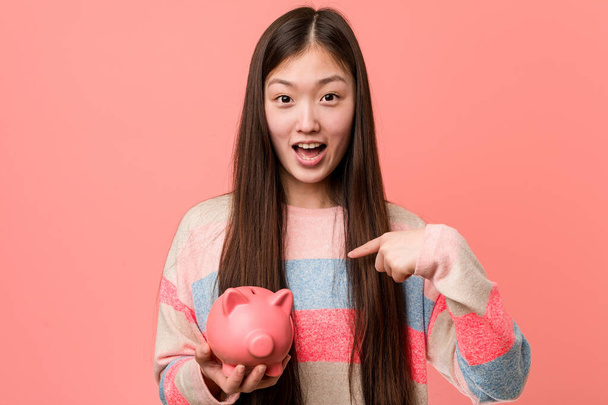 Молода азіатська жінка тримає скарбничку здивований вказуючи на себе, посміхаючись широко
. - Фото, зображення