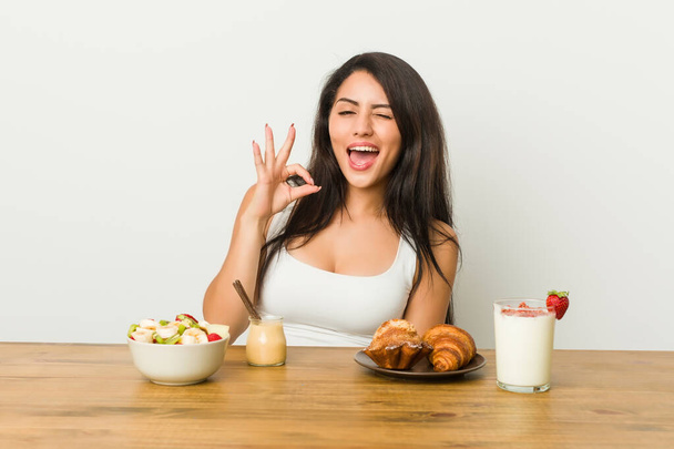 Joven mujer con curvas tomando un desayuno guiña un ojo y sostiene un gesto bien con la mano
. - Foto, Imagen
