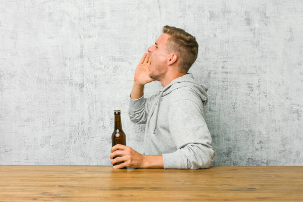 junger Mann trinkt ein Bier auf einem Tisch, schreit und hält Handfläche in der Nähe geöffneten Mundes. - Foto, Bild