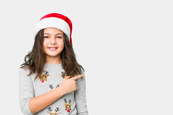 Klein meisje viert kerstdag glimlachend en terzijde wijzend, iets tonend op lege ruimte. - Foto, afbeelding