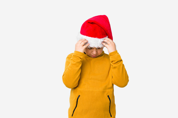 Kleine jongen viert kerst dag het dragen van een kerstman hoed geïsoleerd aanraken tempels en het hebben van hoofdpijn. - Foto, afbeelding