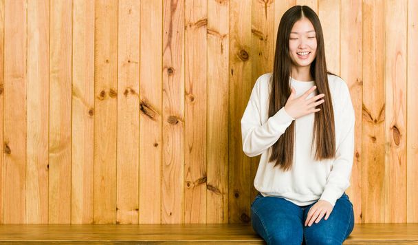 Jovem chinesa sentada em um lugar de madeira ri em voz alta mantendo a mão no peito
. - Foto, Imagem