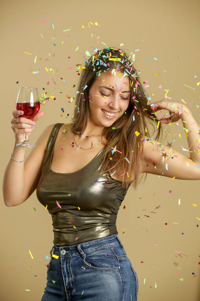 Vonzó nő arany ingben táncol és egy pohár vörösbort tart a kezében. Boldog fiatal és gyönyörű nő élvezi a party konfettivel. - Fotó, kép