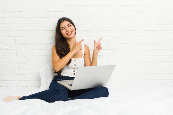 Mujer árabe joven que trabaja con su portátil en la cama apuntando con los dedos índice a un espacio de copia, expresando emoción y deseo
. - Foto, Imagen