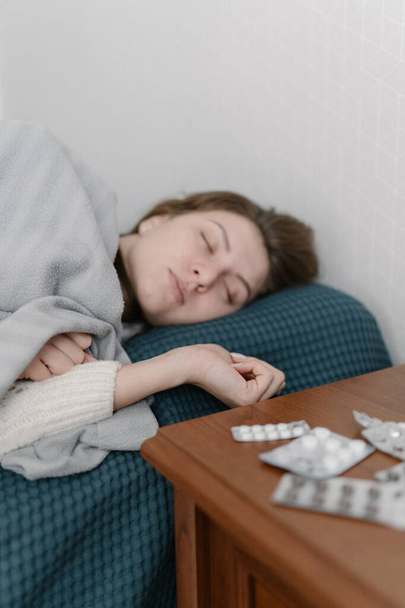 Γυναικείο πορτρέτο στο κρεβάτι - Ailing Καυκάσια γυναίκα σε αναρρωτική άδεια βρίσκεται και κοιμούνται σε ένα λευκό πουλόβερ στο κρεβάτι στο υπνοδωμάτιο. Γρίπη και κρύος ιός στην εποχή των ασθενειών - Φωτογραφία, εικόνα
