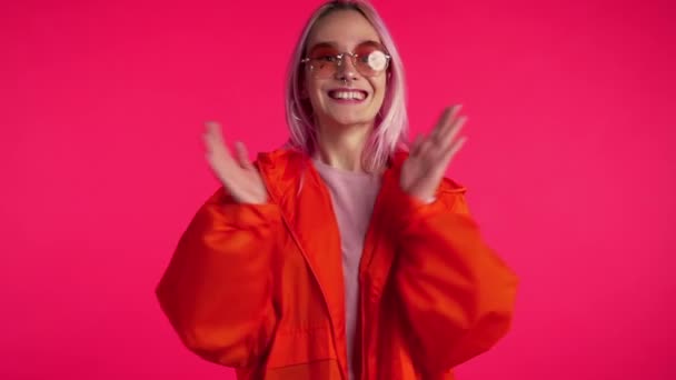 Portrait of girl with dyed pink hair, she claps her hands with delight. Surprised excited happy woman. Mixed race female shocked model on red background.  - Кадри, відео
