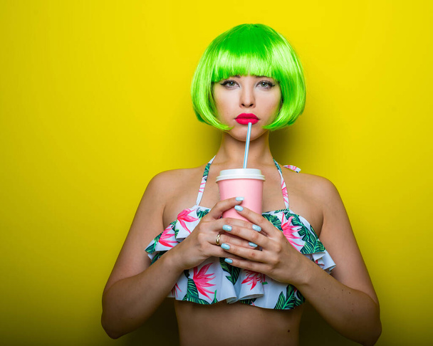 Beautiful young woman in a bright green wig under a quack and a swimsuit posing on a yellow background. Attractive girl with sensual lips flirts on the camera, drinks a drink through a straw. - Photo, Image