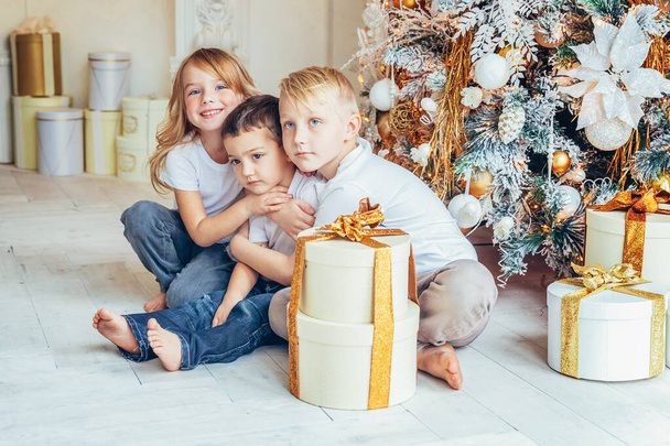 Happy family three children boys and girl relax playing near Christmas tree on Christmas eve at home. Brothers and sister in light bedroom with winter decoration. Christmas New Year time for - Foto, Bild