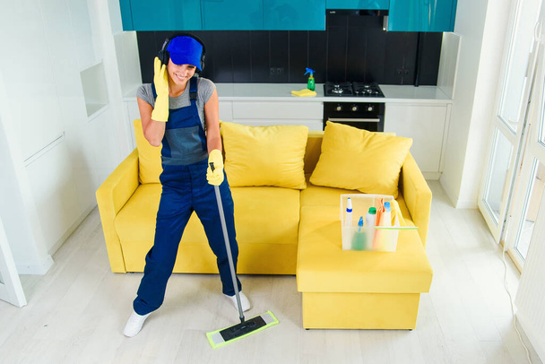 Mujer caucásica como limpiadora profesional en auriculares limpiando piso con fregona y escucha música en casa
. - Foto, Imagen