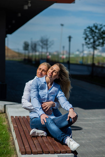 Two surprisingly similar women are sitting on a bench in an embrace and smiling. A middle-aged woman and her elderly mother. Beautiful well-groomed mother and daughter family look. - Photo, Image