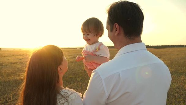 Ο μπαμπάς και η μαμά παίζουν με μια μικρή κόρη στην αγκαλιά της το ηλιοβασίλεμα. οικογενειακές βόλτες με ένα παιδί κατά τη δύση του ηλίου. πατέρας να παίζει με τις κόρες του να ξεκουράζονται στο πάρκο. έννοια της ευτυχης οικογένειας και της παιδικής ηλικίας. - Φωτογραφία, εικόνα