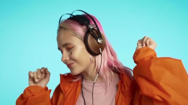 Hipster woman with extraordinary appearance in headphones in orange raincoat on blue background. Pretty girl with pink hair enjoys listening to music.  - Imágenes, Vídeo