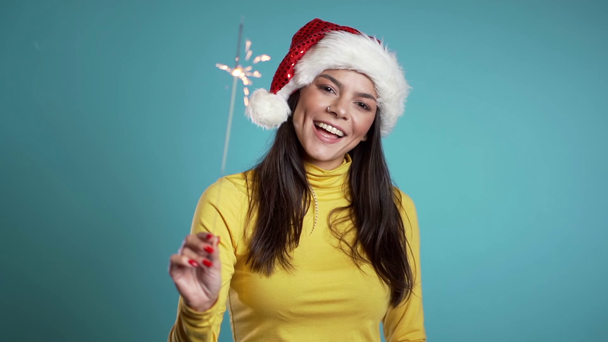 Hispanic girl in Santa hat celebrating with sparkler and bengal fire. Happy mix race woman smiling and having fun against blue background. Slow motion. - Metraje, vídeo