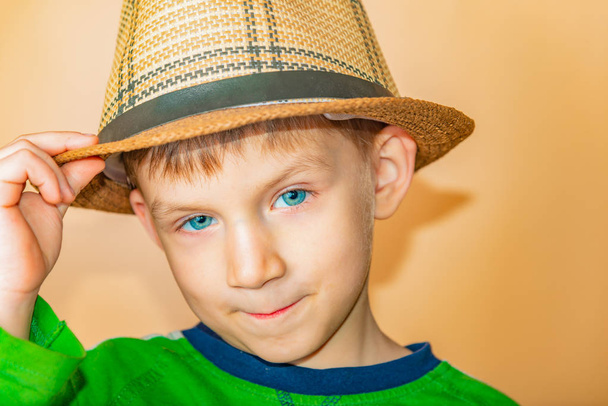 Un niño con un sombrero de paja y ropa verde sobre un fondo beige se quita el sombrero en saludo
. - Foto, imagen