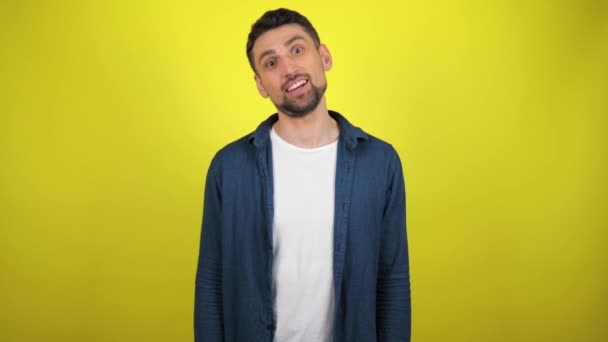 A young man in a blue shirt and white T-shirt is looking to the camera, nods his head and winks. Yellow background with copy space. 4k slowmotion footage - Footage, Video