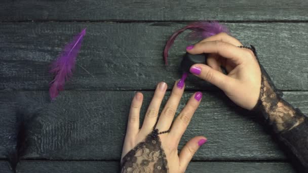 Dziewczyna maluje paznokcie purpurowym żelowym lakierem na czarnym drewnianym tle wśród fioletowych piór. Pełne wdzięku ręce z pięknym manicure na paznokciach.  - Materiał filmowy, wideo