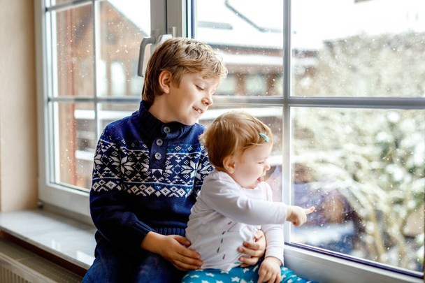Happy adorable kid boy and cute baby girl sitting near window and looking outside on snow on Christmas day or morning. Smiling children, siblings, little sister and brother looking on winter snowfall. - Foto, Imagen