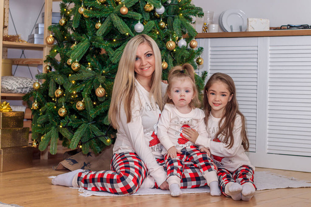 Μαμά και κόρες στο χριστουγεννιάτικο δέντρο. Χριστούγεννα. Αδερφές. Καλά Χριστούγεννα και καλές γιορτές. Χαρούμενη μαμά και οι χαριτωμένες κόρες της ανταλλάσσουν δώρα..  - Φωτογραφία, εικόνα