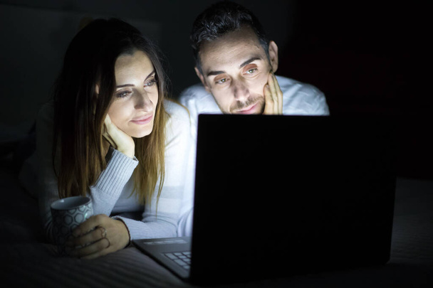 Portrait of a cheerful young couple using laptop computer while laying on bed at night time - Photo, Image
