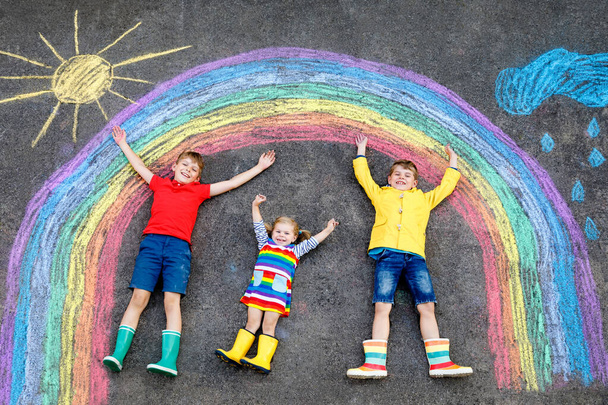 üç küçük çocuk, iki okul çocuk erkek ve yürümeye başlayan kız asfalt üzerinde renkli tebeşir ile gökkuşağı resim çizim ile eğleniyor. Birlikte oynarken yere boyama lastik çizmeler Kardeşler. - Fotoğraf, Görsel