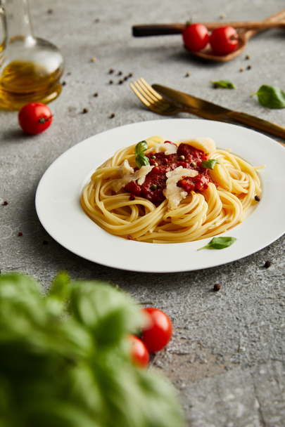 selektivní zaměření chutných boloňských těstovin s rajčatovou omáčkou a parmezánem na bílém talíři v blízkosti ingrediencí a příborů na šedém pozadí - Fotografie, Obrázek