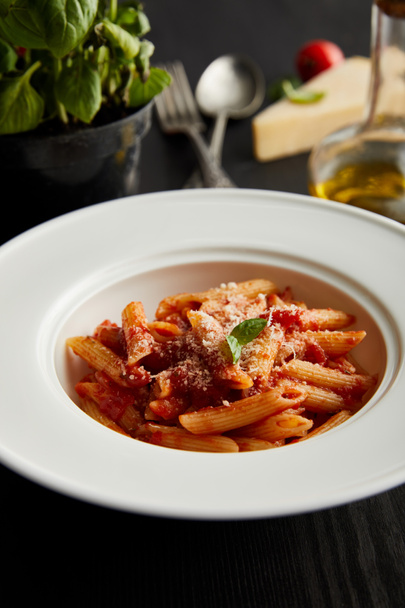 selective focus of tasty bolognese pasta with tomato sauce and Parmesan on white plate near ingredients and cutlery on black wooden background - Photo, Image