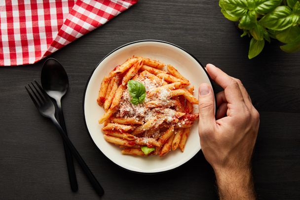 cropped view of man holding plate with tasty bolognese pasta on black wooden table with basil, cutlery and check napkin - 写真・画像