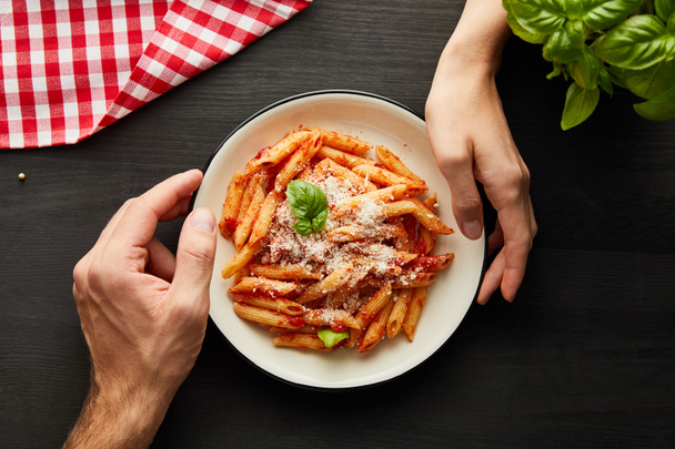 cropped view of couple holding plate with tasty bolognese pasta on black wooden table with basil, cutlery and check napkin - Photo, Image