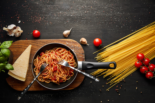 top view of tasty bolognese pasta in frying pan near ingredients and cutlery on black wooden background - Photo, Image