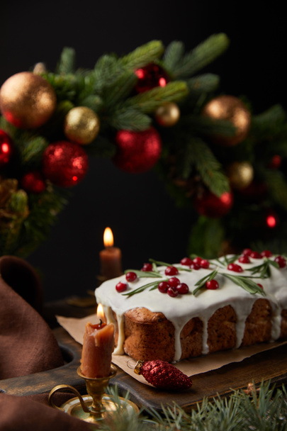 selektivní zaměření tradičního vánočního dortu s brusinkami u vánočního věnce s cetkami a hořícími svíčkami na dřevěném stole izolovaném na černém - Fotografie, Obrázek