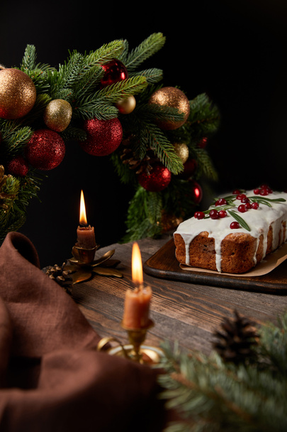 selective focus of traditional Christmas cake with cranberry near Christmas wreath with baubles and burning candles on wooden table isolated on black - Photo, Image
