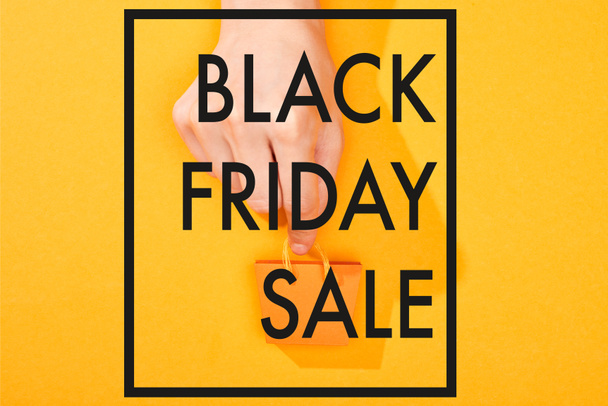 cropped view of hand holding small shopping bag on bright orange background with black Friday sale illustration - Foto, imagen