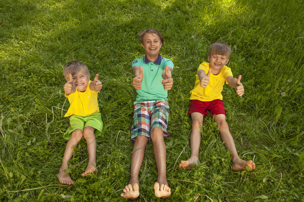 Drie lachende jongens zitten op groen gras en tonen duimen omhoog. - Foto, afbeelding