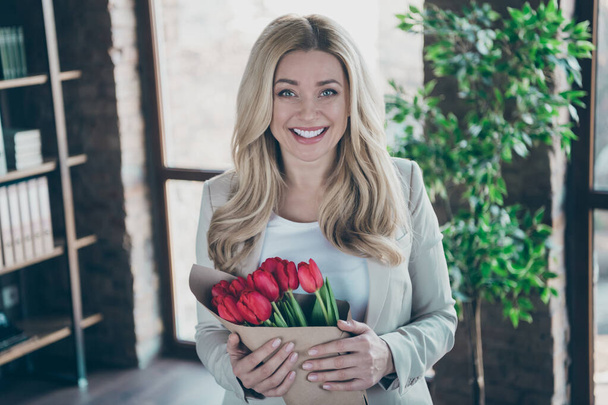 Photo of beautiful blond business lady came workplace morning holding fresh tulips bunch in arms overjoyed husband surprise modern comfortable office - Фото, изображение