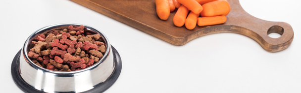 comida seca para mascotas en un tazón cerca de zanahoria cruda en una tabla de cortar de madera aislada en blanco, tiro panorámico
 - Foto, imagen