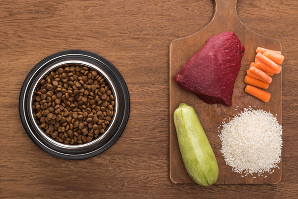 Draufsicht auf Trockenfutter in silberner Schüssel in der Nähe von rohem Fleisch, Zucchini, Reis und Karotten auf Holztisch - Foto, Bild