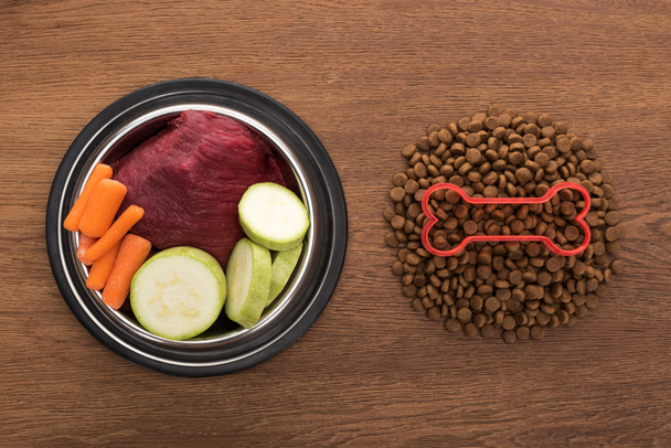 vista superior de alimentos secos para mascotas con hueso cerca de verduras crudas y carne en un tazón sobre una mesa de madera
 - Foto, Imagen